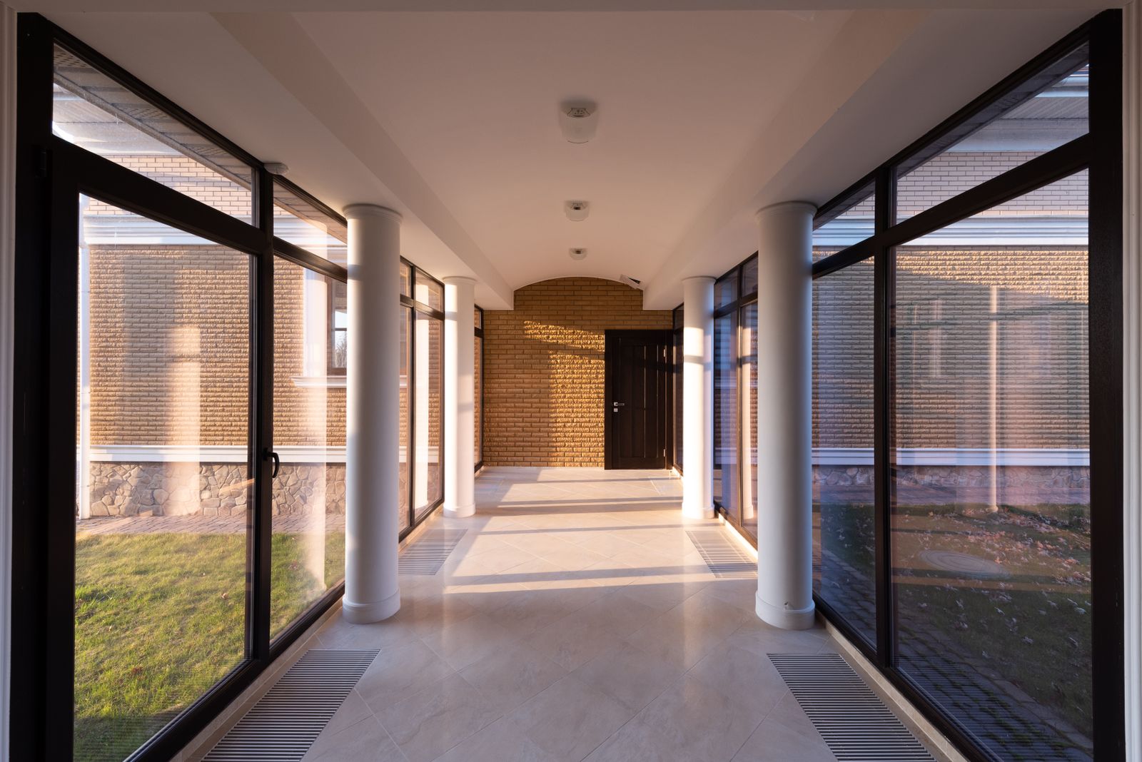 After cleaning: Empty long hallway with columns leading to brick wall and door between glass transparent walls of modern building overlooking grassy lawn